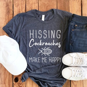 May include: A dark gray t-shirt with the text "HISSING Cockroaches MAKE ME HAPPY" in white. A white cockroach illustration is above the text. The shirt is laid out on a wooden surface with a white baseball cap, blue jeans, and white sneakers.