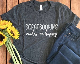 Può includere: Una maglietta grigia scura con il testo "Scrapbooking makes me happy" in lettere bianche. La maglietta è appoggiata su una superficie di legno con un paio di jeans blu e un mazzo di girasoli gialli.