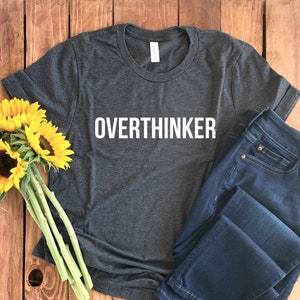 May include: A dark gray t-shirt with the word "Overthinker" printed in white on the front. The shirt is laid flat on a wooden surface with a bouquet of sunflowers and a pair of blue jeans.