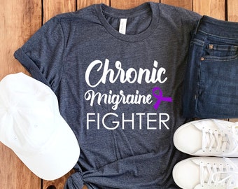 May include: A dark gray t-shirt with the text "Chronic Migraine Fighter" in white lettering. A purple ribbon is next to the text. The shirt is laid out on a wooden surface with a white baseball cap, a pair of blue jeans, and a pair of white sneakers.