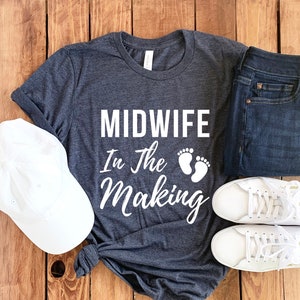 May include: A dark grey t-shirt with the text "Midwife in the Making" and a pair of white footprints. The shirt is laid out on a wooden surface with a white baseball cap, a pair of blue jeans, and two white trainers.
