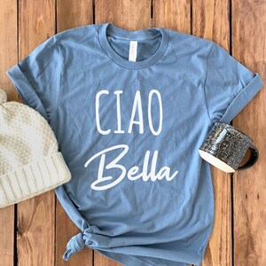 May include: A blue t-shirt with the text "Ciao Bella" in white lettering. The shirt is on a wooden surface with a beige knitted hat and a black mug.