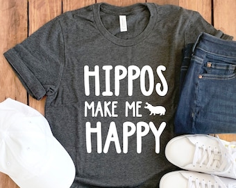 May include: A dark gray t-shirt with white text that reads "Hippos make me happy" and a small white hippo illustration. The shirt is laid out on a wooden surface with a white baseball cap, blue jeans, and white sneakers.
