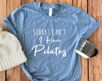 May include: A blue t-shirt with the text "Sorry I can't I have Pilates" in white lettering. The shirt is on a wooden surface with a white knit hat and a black mug.