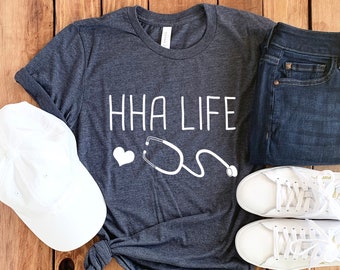 Peut inclure: Un t-shirt gris foncé avec le texte "HHA LIFE" en lettres blanches. Un cœur blanc et un stéthoscope sont également imprimés sur le t-shirt. Le t-shirt est posé sur une surface en bois avec une casquette de baseball blanche, un jean bleu et une paire de baskets blanches.