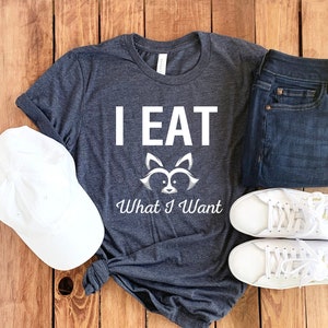 May include: A dark gray t-shirt with white text that reads "I EAT What I Want" and a raccoon graphic. The shirt is paired with white sneakers, blue jeans, and a white baseball cap.