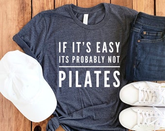 Peut inclure: Un t-shirt gris foncé avec un texte blanc qui dit "Si c'est facile, ce n'est probablement pas du Pilates". Le t-shirt est posé sur une surface en bois avec une casquette de baseball blanche, un jean bleu et deux baskets blanches.
