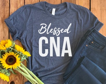 May include: A dark blue t-shirt with the text "Blessed CNA" in white lettering. The shirt is laying on a wooden surface with a bouquet of sunflowers.