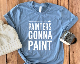 May include: A pale blue t-shirt with the white text "PAINTERS GONNA PAINT" and a paintbrush graphic. A cream-coloured knitted beanie and a black mug with a white interior and a pattern of faces are also present. The items are on a wooden surface.