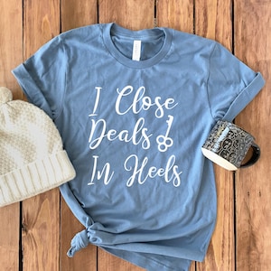 May include: A blue t-shirt with white text that reads "I Close Deals In Heels" with a key and a keyhole graphic. The shirt is on a wooden surface with a white knit beanie and a black mug.