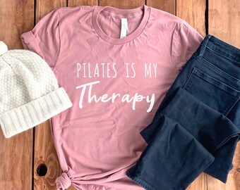 Peut inclure: Un t-shirt rose avec le texte "Pilates is my Therapy" en lettres blanches. Le t-shirt est associé à un jean bleu et un bonnet en tricot blanc.