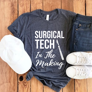 May include: A dark gray t-shirt with the text "Surgical Tech In the Making" and a white scalpel graphic. The shirt is laid out on a wooden surface with a white baseball cap, blue jeans, and white sneakers.