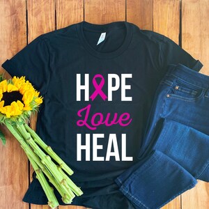 Puede incluir: Camiseta negra con una cinta rosa, las palabras "HOPE", "Love" y "HEAL" en texto blanco.