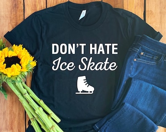 May include: Black t-shirt with white text that reads "Don't Hate Ice Skate" and a white illustration of an ice skate. The t-shirt is laying on a wooden surface with a pair of blue jeans.