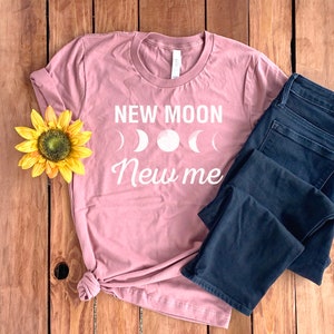 Peut inclure: Un t-shirt rose poussi&eacute;reux avec le texte "NEW MOON New me" en lettres blanches. Le texte est accompagn&eacute; d'un graphique de quatre croissants de lune blancs.