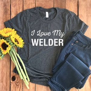 May include: A dark gray t-shirt with the text "I Love My Welder" in white, paired with dark blue jeans and a bouquet of sunflowers. The shirt is a crew neck style.