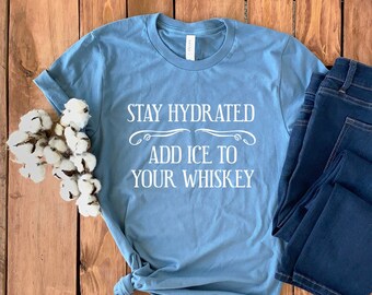 May include: A light blue t-shirt with the text "Stay Hydrated Add Ice To Your Whiskey" in white. The shirt is laid out on a wooden surface with a pair of blue jeans.