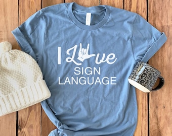 May include: A blue t-shirt with the text "I Love Sign Language" in white. The text includes a hand sign for "love". The shirt is on a wooden surface with a white knit beanie and a black mug.