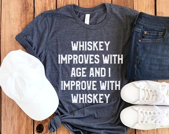May include: A dark gray t-shirt with the text "Whiskey improves with age and I improve with whiskey" printed in white. The shirt is laid out on a wooden surface with a white baseball cap, blue jeans, and white sneakers.