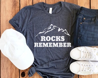May include: A dark grey t-shirt with a white mountain graphic and the text "Rocks Remember". The shirt is laid out on a wooden surface with a white baseball cap, a pair of blue jeans, and two white trainers.