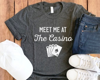 Könnte beinhalten: Ein dunkelgraues T-Shirt mit dem Text "Meet me at The Casino" und einer Grafik von vier Spielkarten. Das Shirt wird mit einer blauen Jeans, weißen Turnschuhen und einer weißen Baseballkappe kombiniert.