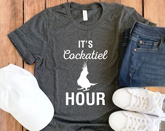 May include: A dark gray t-shirt with the text "It's Cockatiel Hour" in white. A white cockatiel silhouette is in the center of the shirt. The shirt is laid out on a wooden surface with a white baseball cap, blue jeans, and white sneakers.