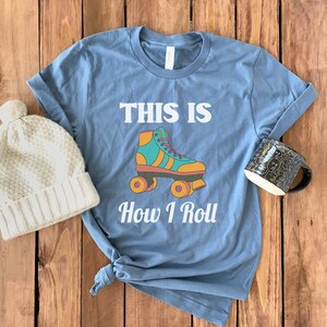 K&ouml;nnte beinhalten: Ein blaues T-Shirt mit dem Text "This is How I Roll" und einer Grafik eines Rollschuhs. Der Rollschuh ist gelb, orange, gr&uuml;n und blau.