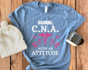 May include: A blue t-shirt with the text "WARNING: C.N.A. WITH AN ATTITUDE" in white and a pink caduceus symbol.