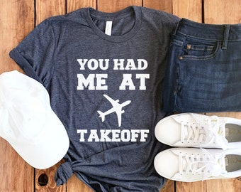 May include: A dark gray t-shirt with the text "You had me at takeoff" and a white airplane graphic. The shirt is paired with white sneakers, blue jeans, and a white baseball cap.