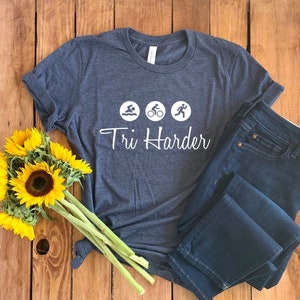 K&ouml;nnte beinhalten: Ein dunkelblaues T-Shirt mit wei&szlig;em Text, der "Tri Harder" lautet, und drei Symbolen, die Schwimmen, Radfahren und Laufen darstellen.