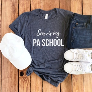 Op de afbeelding: Een donkergrijs T-shirt met de tekst "Surviving PA School" in witte letters. Het shirt ligt op een houten oppervlak met een witte baseballpet, een blauwe spijkerbroek en twee witte sneakers.