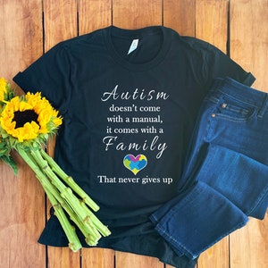 Op de afbeelding: Zwart T-shirt met witte tekst die luidt: "Autisme komt niet met een handleiding, het komt met een familie die nooit opgeeft". Een kleurrijk puzzelhart bevindt zich in het midden van het woord "Familie". Het shirt ligt op een houten oppervlak met een blauwe jeans en zonnebloemen.