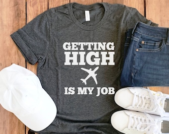 May include: A dark gray t-shirt with the text "GETTING HIGH IS MY JOB" in white lettering and a white airplane graphic. The shirt is paired with blue jeans, white sneakers, and a white baseball cap.