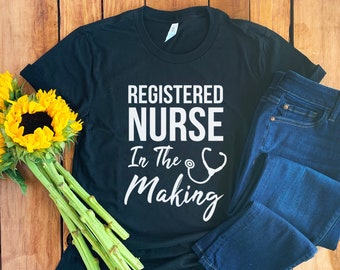 Könnte beinhalten: Schwarzes T-Shirt mit weißem Text, der "Registered Nurse In The Making" lautet, mit einem Stethoskop-Motiv. Das Shirt liegt auf einer Holzoberfläche mit einer blauen Jeans und einem Sonnenblumenstrauß.