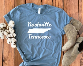 May include: A dusty blue t-shirt with the words "Nashville Tennessee" and a white outline of the state of Tennessee. The shirt is knotted at the bottom and paired with a pair of gray ankle boots. Cotton stems are placed on the left side.