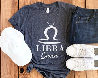 May include: A dark gray t-shirt with the text "LIBRA Queen" and a Libra symbol with a crown. The shirt is paired with blue jeans, white sneakers, and a white baseball cap. The items are arranged on a wooden surface.