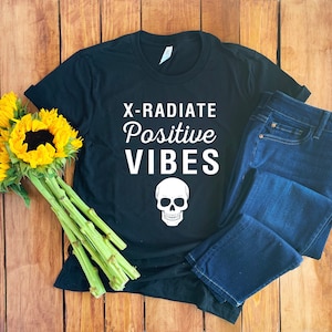 Op de afbeelding: Zwart T-shirt met witte tekst die "X-RADIATE Positive VIBES" zegt en een witte schedel afbeelding. Het shirt ligt op een houten oppervlak met een blauwe jeans.