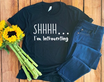May include: Black t-shirt with white text that reads "SHHHH... I'm Introverting". The shirt is laid out on a wooden surface with a bouquet of sunflowers and a pair of blue jeans.