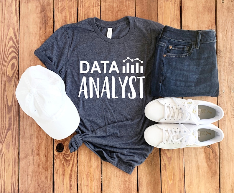 May include: A dark gray t-shirt with the text "DATA ANALYST" in white lettering and a white graph icon. The shirt is laid out on a wooden surface with a white baseball cap, a pair of blue jeans, and two white sneakers.