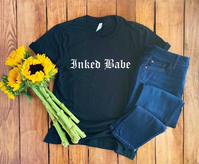 May include: Black t-shirt with the text "Inked Babe" in white gothic lettering. The shirt is laid out on a wooden surface with a pair of blue jeans and a bouquet of sunflowers.