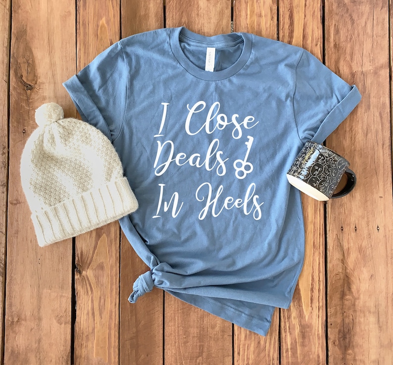 May include: A blue t-shirt with white text that reads "I Close Deals In Heels" with a key and a keyhole graphic. The shirt is on a wooden surface with a white knit beanie and a black mug.