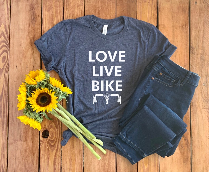 May include: A dark blue t-shirt with the text "LOVE LIVE BIKE" in white lettering. The shirt also features a white silhouette of a bicycle handlebar. The shirt is laid out on a wooden surface with a bouquet of sunflowers.