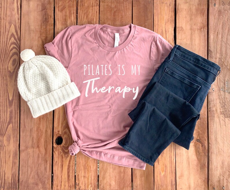 Peut inclure: Un t-shirt rose avec le texte "Pilates is my Therapy" en lettres blanches. Le t-shirt est associ&eacute; &agrave; un jean bleu et un bonnet en tricot blanc.