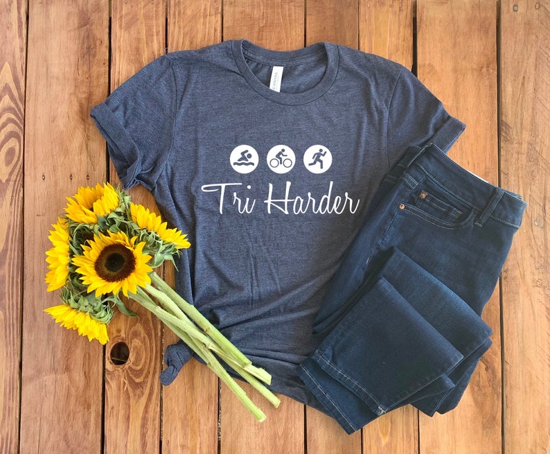 K&ouml;nnte beinhalten: Ein dunkelblaues T-Shirt mit wei&szlig;em Text, der "Tri Harder" lautet, und drei Symbolen, die Schwimmen, Radfahren und Laufen darstellen.