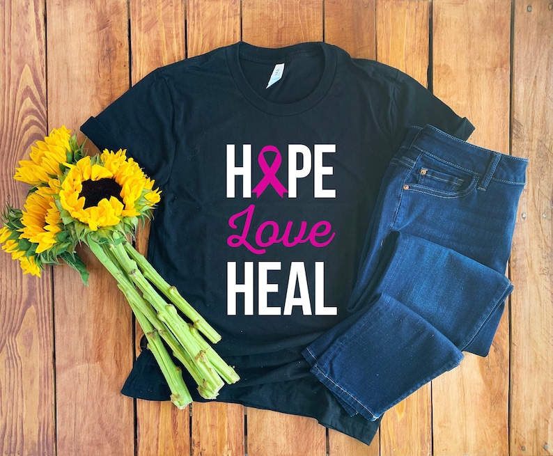 Puede incluir: Camiseta negra con una cinta rosa, las palabras "HOPE", "Love" y "HEAL" en texto blanco.