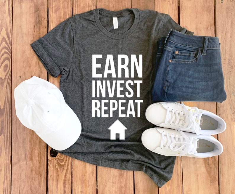 Może przedstawiać: Szary t-shirt z nadrukiem "EARN INVEST REPEAT" w białym kolorze i białym ikoną domu. T-shirt leży na drewnianej powierzchni z parą białych but&oacute;w sportowych, białą czapką z daszkiem i niebieskimi dżinsami.