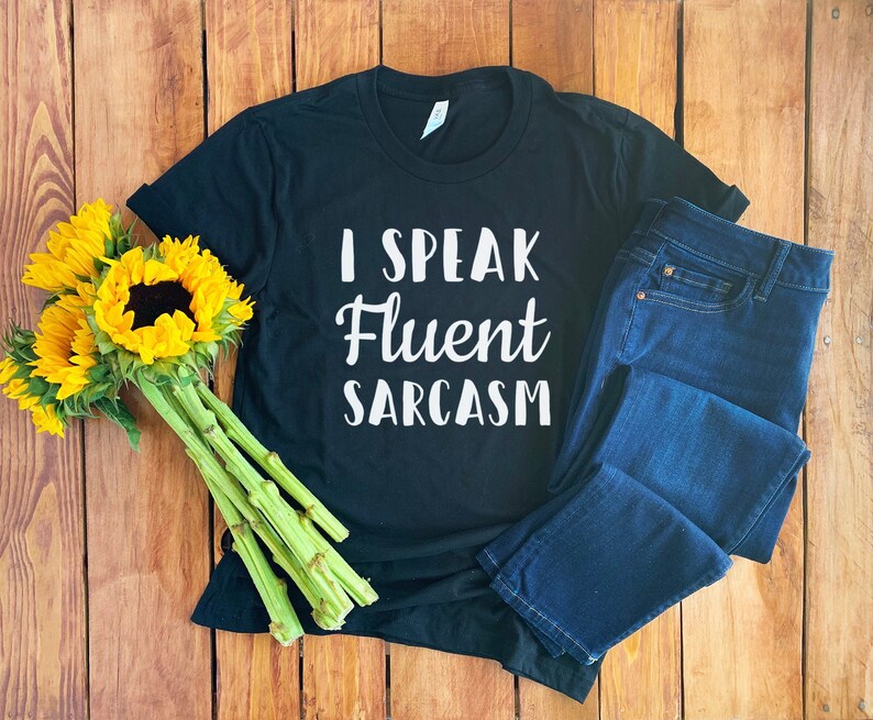 May include: Black t-shirt with white text that reads "I speak fluent sarcasm". The shirt is laying on a wooden surface with a bouquet of sunflowers and a pair of blue jeans.