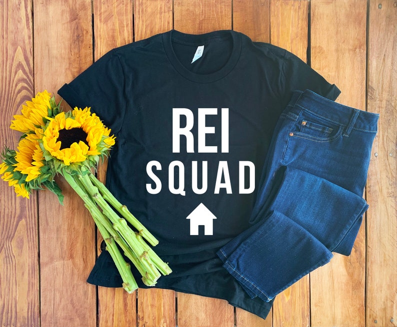 以下が含まれることがあります： 白い文字で「REI SQUAD」と白い家のアイコンが書かれた黒いTシャツ。Tシャツは木製の表面に置かれ、青いジーンズが置かれている。