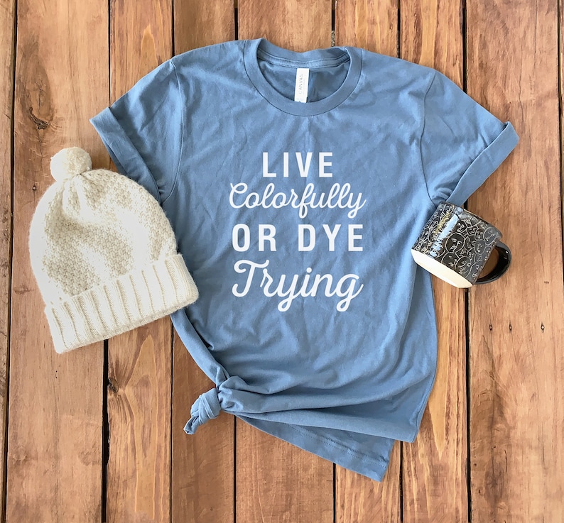 Op de afbeelding: Een blauw t-shirt met de tekst "Live Colorfully Or Dye Trying" in witte letters. Het shirt ligt op een houten oppervlak met een witte gebreide muts en een zwarte mok.