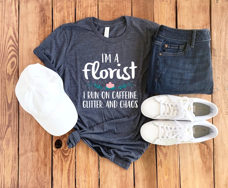 May include: A dark gray t-shirt with the text "I'm a florist I run on caffeine, glitter, and chaos". The shirt is laid out on a wooden surface with a white baseball cap, a pair of blue jeans, and a pair of white sneakers.